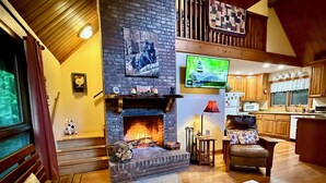A cozy living room with a fireplace, wooden furniture, a television, and a kitchen area.