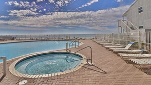 En swimmingpool med et spabad, omgivet af liggestole og en terrassedæk i mursten, under en delvist overskyet himmel.