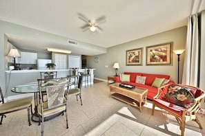 A living room with a red sofa, a glass dining table, and chairs. There is a kitchen in the background with white cabinets and a ceiling fan.