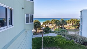 A view of a building with a balcony overlooking a beach, palm trees, and a clear sky. - Boardwalk 4BR/3 Bath **2nd Floor** End Unit (Panama City Beach)