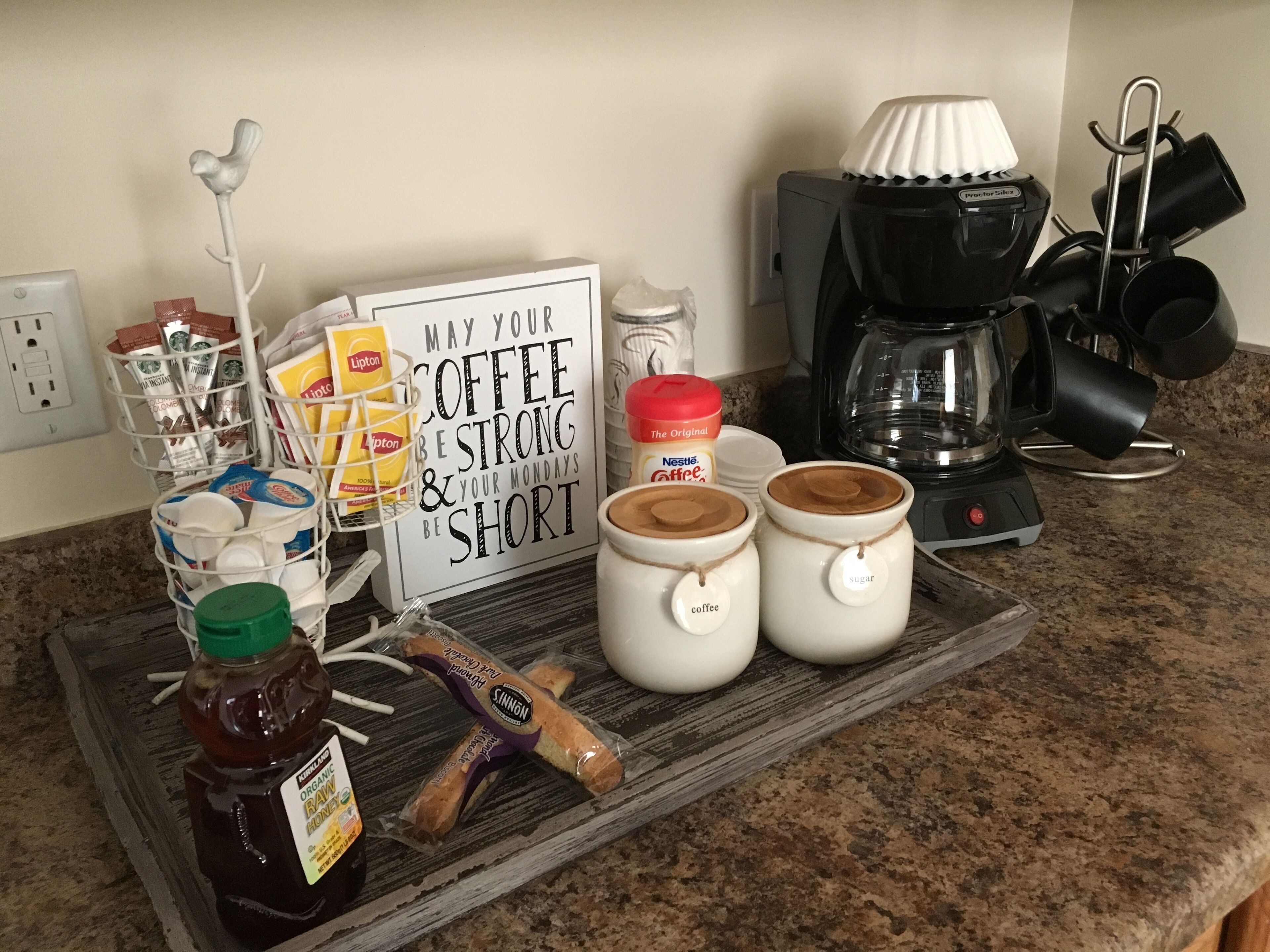 Un comptoir de cuisine avec tout le nécessaire pour préparer du café, notamment une machine à café, des bocaux étiquetés « café » et « sucre », un plateau avec des dosettes de café, un pot de miel et un panneau affichant un message motivant.