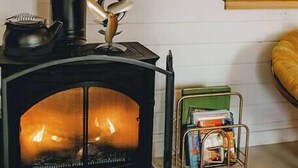 A wood-burning stove with a visible fire, a small shelf with books, and a window with a wooden frame.