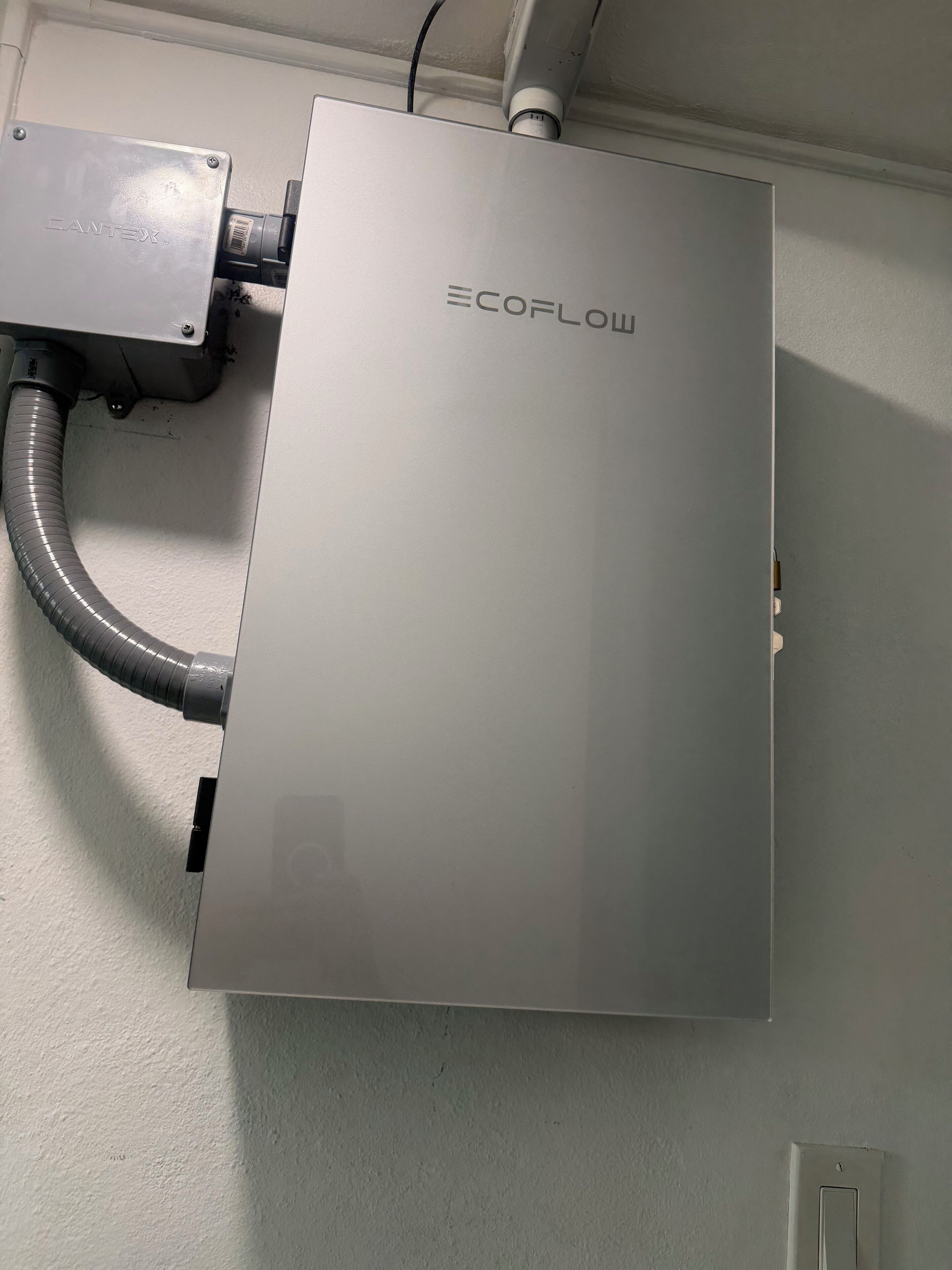 Un appareil métallique rectangulaire portant l’inscription « ECOFLOW », fixé au mur, et auquel est raccordé un conduit flexible.
