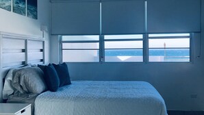 Een slaapkamer met een bed, nachtkastje, dressoir, plafondventilator, airconditioning en een raam met uitzicht op zee.