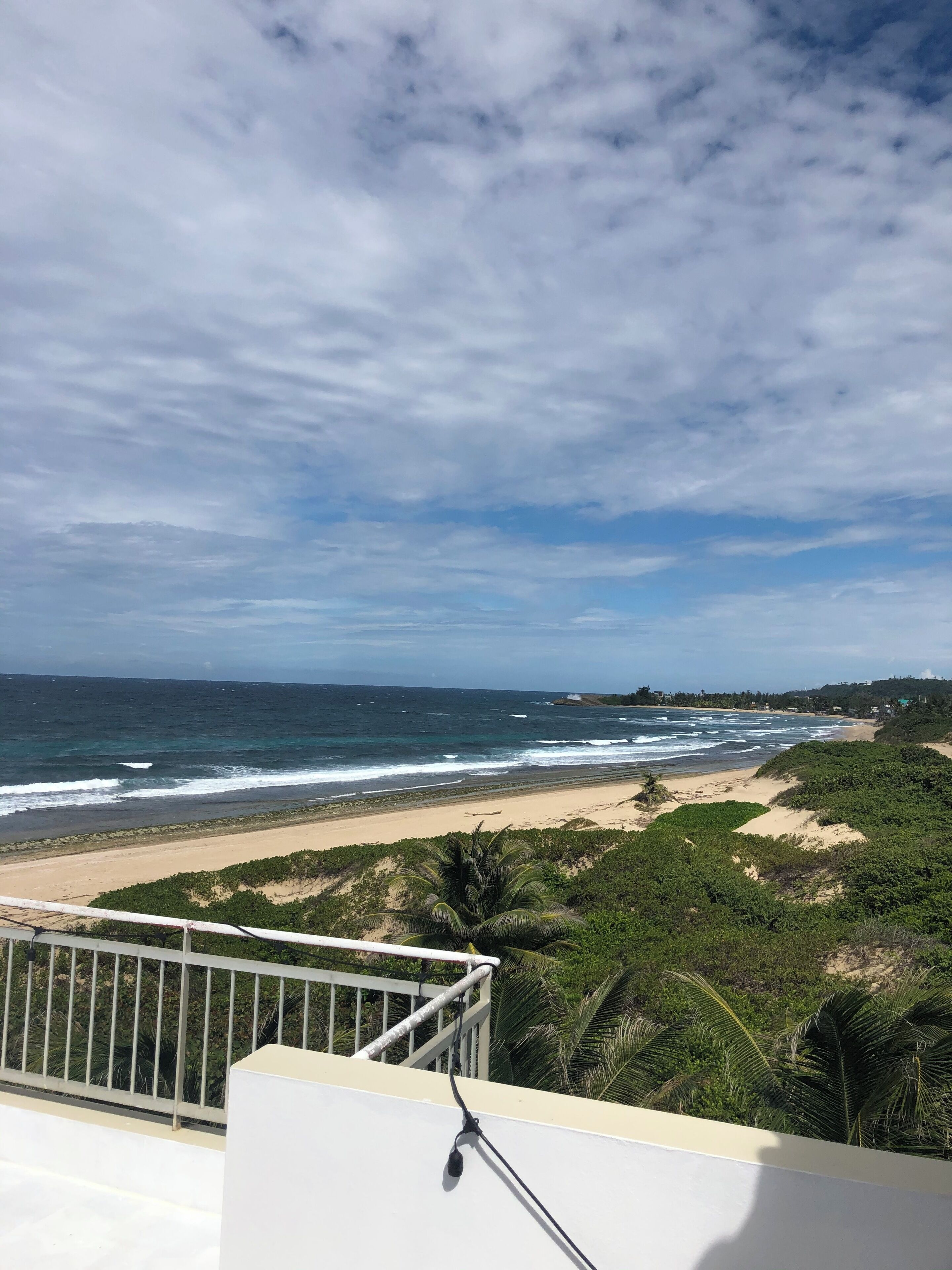 A coastal view from a balcony with a railing, ocean waves, sandy beach, and greenery.