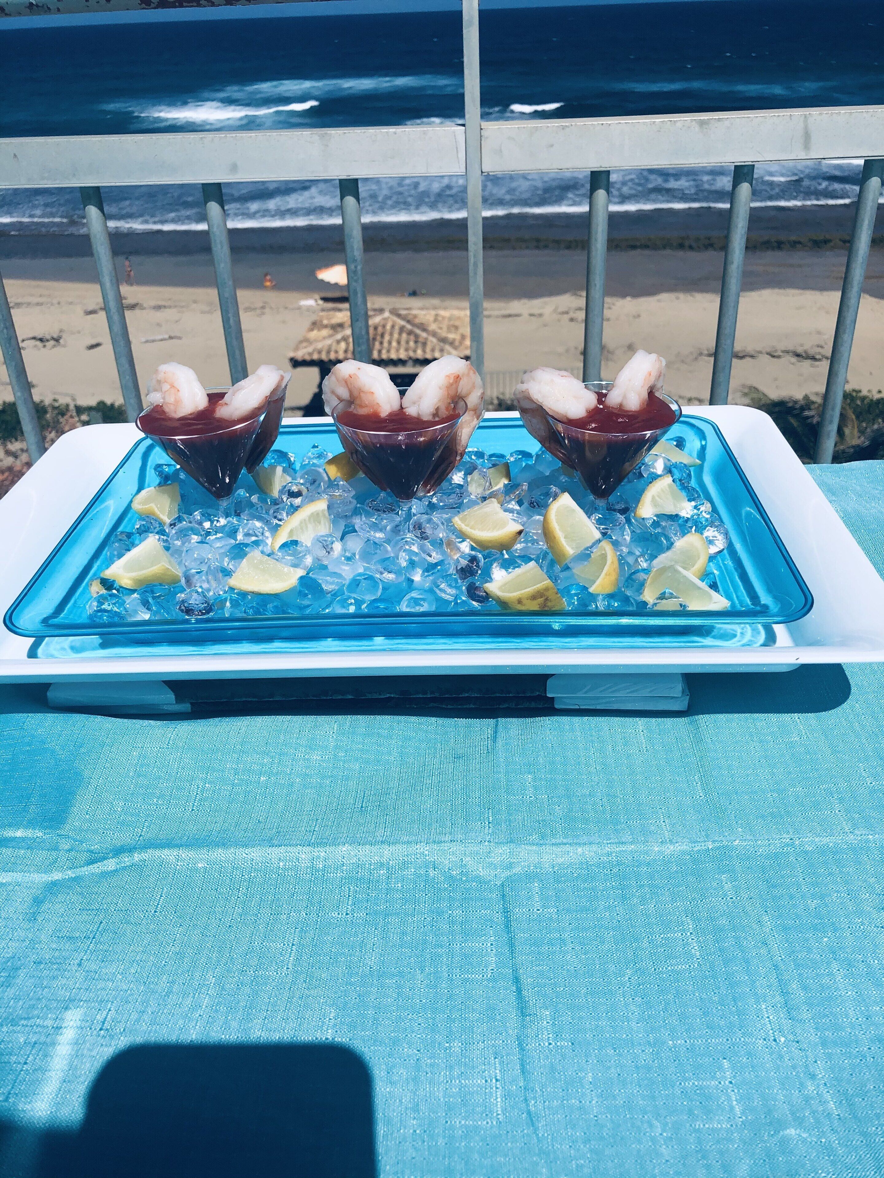A tray with three martini glasses containing seafood cocktails, surrounded by ice cubes and lemon slices, set on a blue table with a view of the ocean and beach in the background.