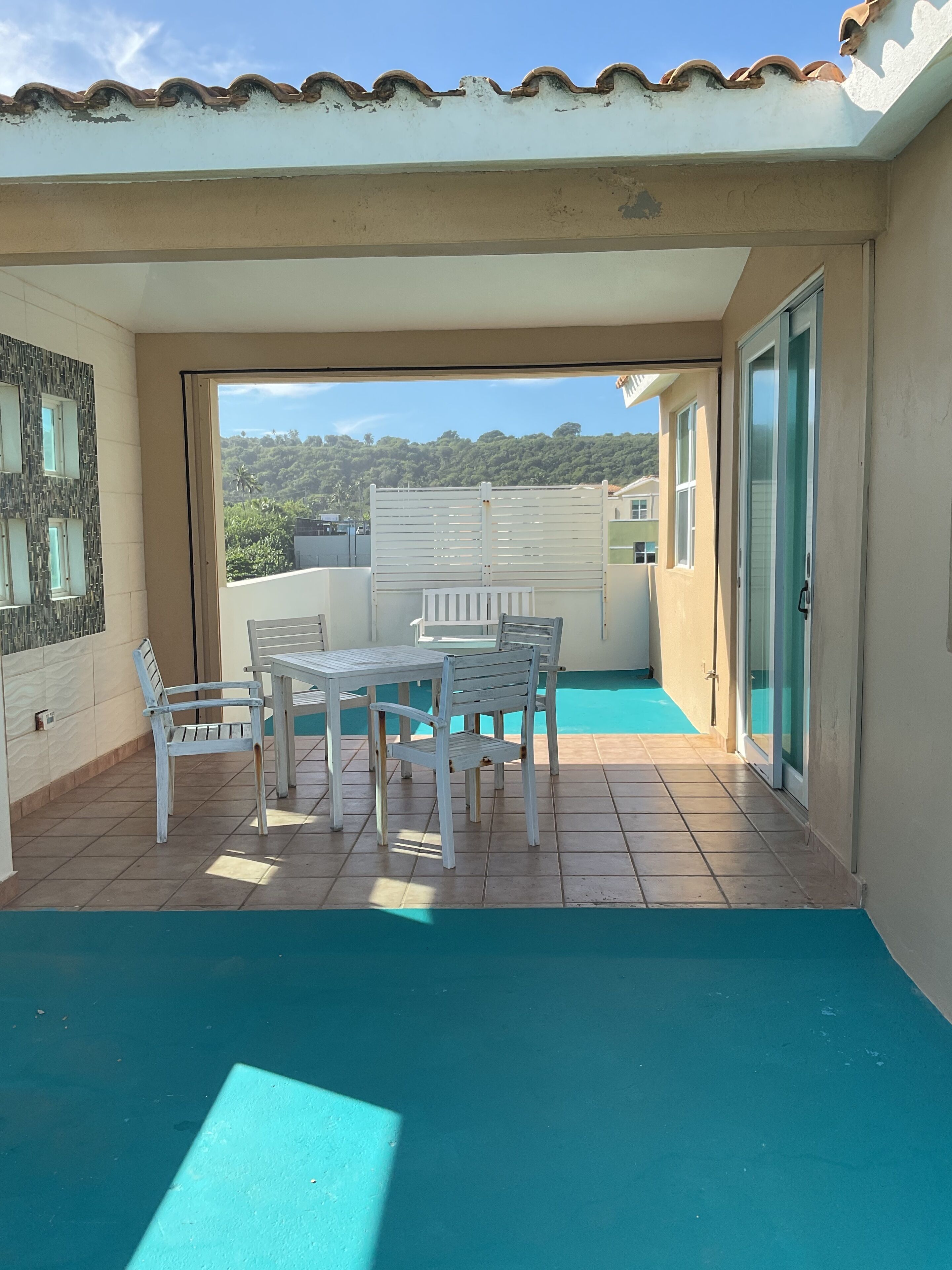 Ein überdachter Außenbereich mit Tisch und Stühlen, Blick auf die umliegende Landschaft und eine Tür zu einem angrenzenden Raum.