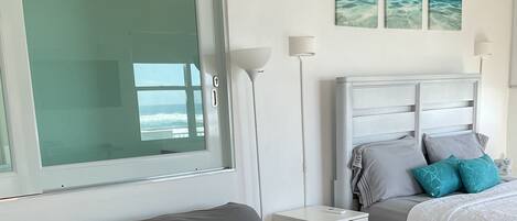 A bedroom with a grey sofa, a bed with a white duvet, and a window with a view of the ocean.