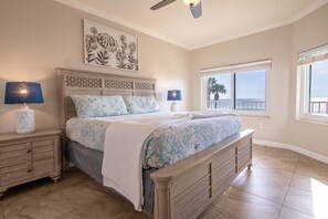 A bedroom with a large bed, two bedside tables, a ceiling fan, a window with a view, and a decorative wall piece.