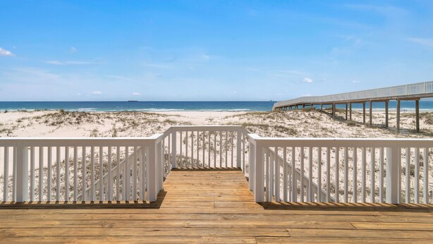 Une terrasse en bois menant à une plage, avec des rampes blanches et une promenade en bois en arrière-plan.
