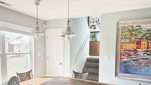 Un espace repas comprenant une grande table, des chaises, des luminaires suspendus, un escalier et un tableau accroché au mur.