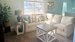 Un salon avec un canapé d’angle beige, une petite table d’appoint avec une lampe, une table basse en verre, une grande plante en pot et une fenêtre avec des stores.