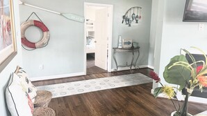Un couloir avec un plancher en bois, une porte blanche donnant sur une chambre, une petite table ornée d’un objet décoratif et une plante en pot.