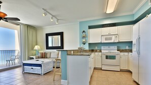 A compact kitchen with white cabinets, a microwave, and a refrigerator. There is a dining area with a table and chairs, and a balcony with a view. - Tidewater 1011 10th Flr 2BR+Bunks 3BA Early Booking Discounts 2 Beach Chairs (Panama City Beach)
