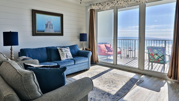 A living room with a blue sofa, a grey armchair, a ceiling fan, and a large window with a view of the sea. - 1st floor 3br Marisol unit sleeps 8! (Panama City Beach)