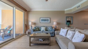 A living room with a sofa, a coffee table, and chairs. There is a large window and a door leading to a balcony.