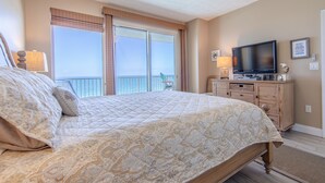 A bedroom with a large bed, a television, a dresser, and a view of the ocean through the sliding doors.