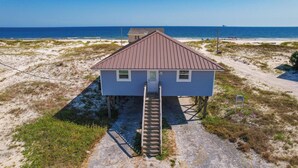 A small, elevated wooden structure with a metal roof, a wooden staircase leading up to a door, and a clear view of the beach and ocean in the background.