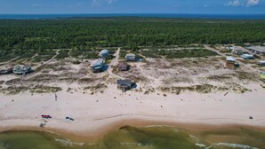 An aerial view of a coastal area with several buildings, including a large house with multiple rooms and a smaller structure, surrounded by a forested area and a sandy beach.
