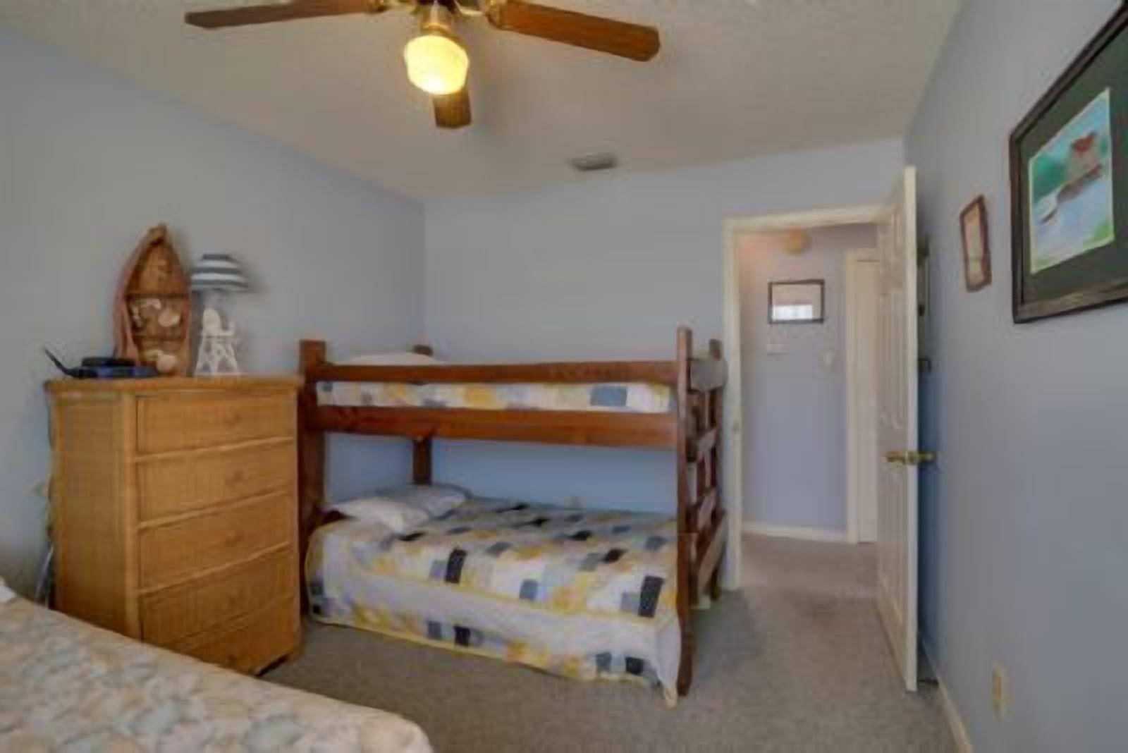 Un dormitorio con literas, un aparador de madera, un ventilador de techo y una puerta que da a otra habitación.