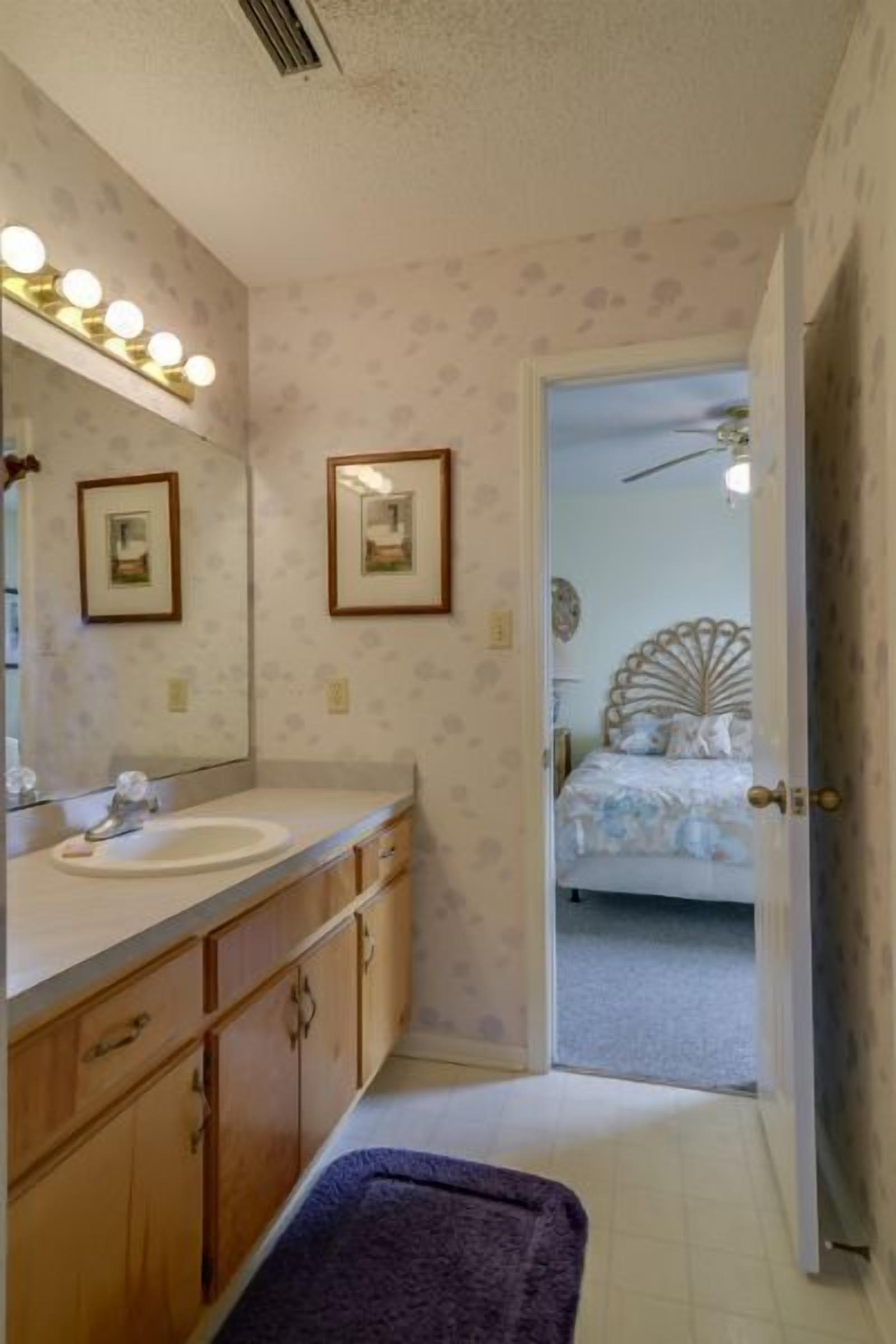 Une salle de bain avec un meuble-lavabo, un miroir et des luminaires. Une porte donne accès à une chambre à coucher équipée d’un lit et d’un ventilateur de plafond.