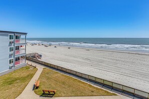 Un hotel sulla spiaggia con un balcone, una spiaggia sabbiosa e una recinzione in legno.