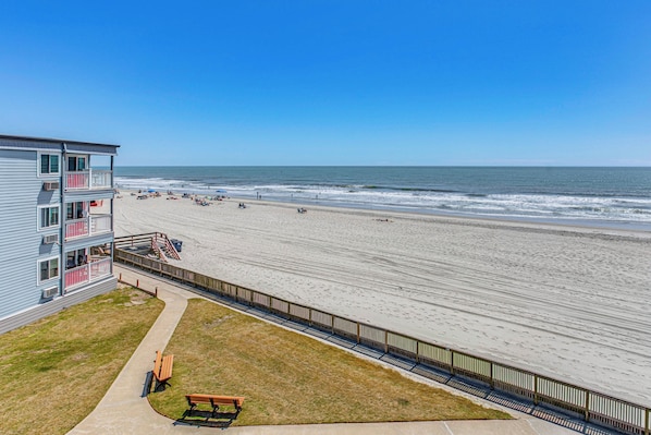Ein Strandhotel mit Balkon, Sandstrand und Holzzaun.