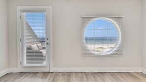 A room with a door leading to a balcony, a window with a view of the beach, and a light-colored wooden floor.