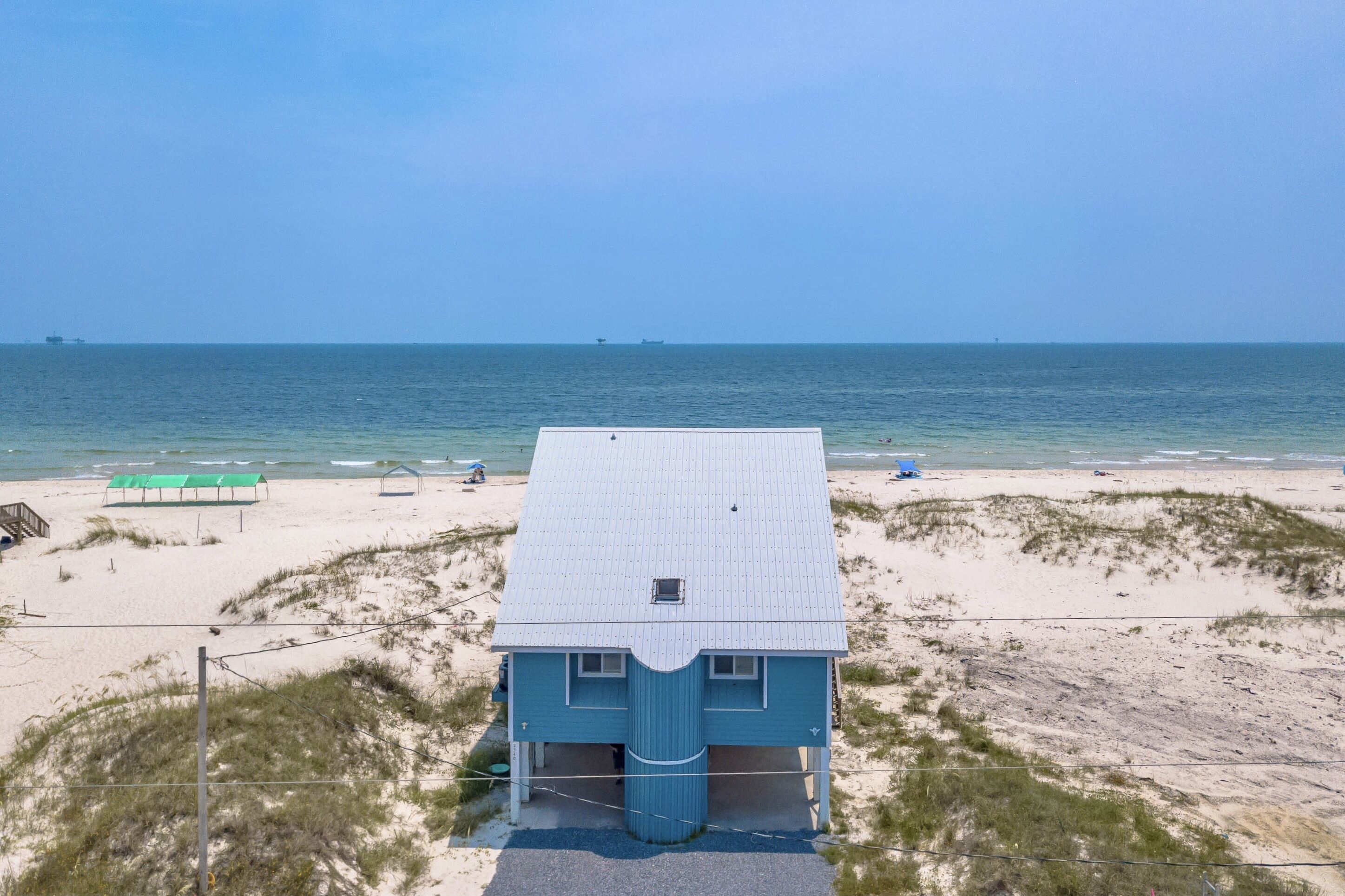 Une maison de plage avec un toit blanc et une façade bleue, située sur une plage de sable fin, avec des dunes en premier plan.