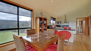 A modern kitchen with a large window, a dining area with a glass table, and a view of a green landscape. - Moon River Retreat - Panoramic water views (Gardners Bay)