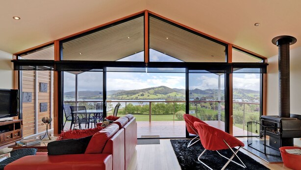 A spacious living room with a large glass wall offering a scenic view of hills and a lake. - Moon River Retreat - Panoramic water views (Gardners Bay)