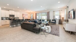 A modern living room with a sectional sofa, a dining area with a kitchen in the background, and large windows providing natural light. - Modern 4 Bedroom Home with Game Room in Encore Resort (Kissimmee)