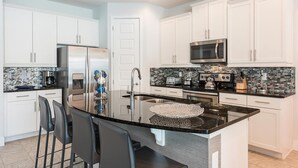 A modern kitchen with white cabinets, a stainless steel refrigerator, a microwave, and a large island with a sink. - Modern 4 Bedroom Home with Game Room in Encore Resort (Kissimmee)