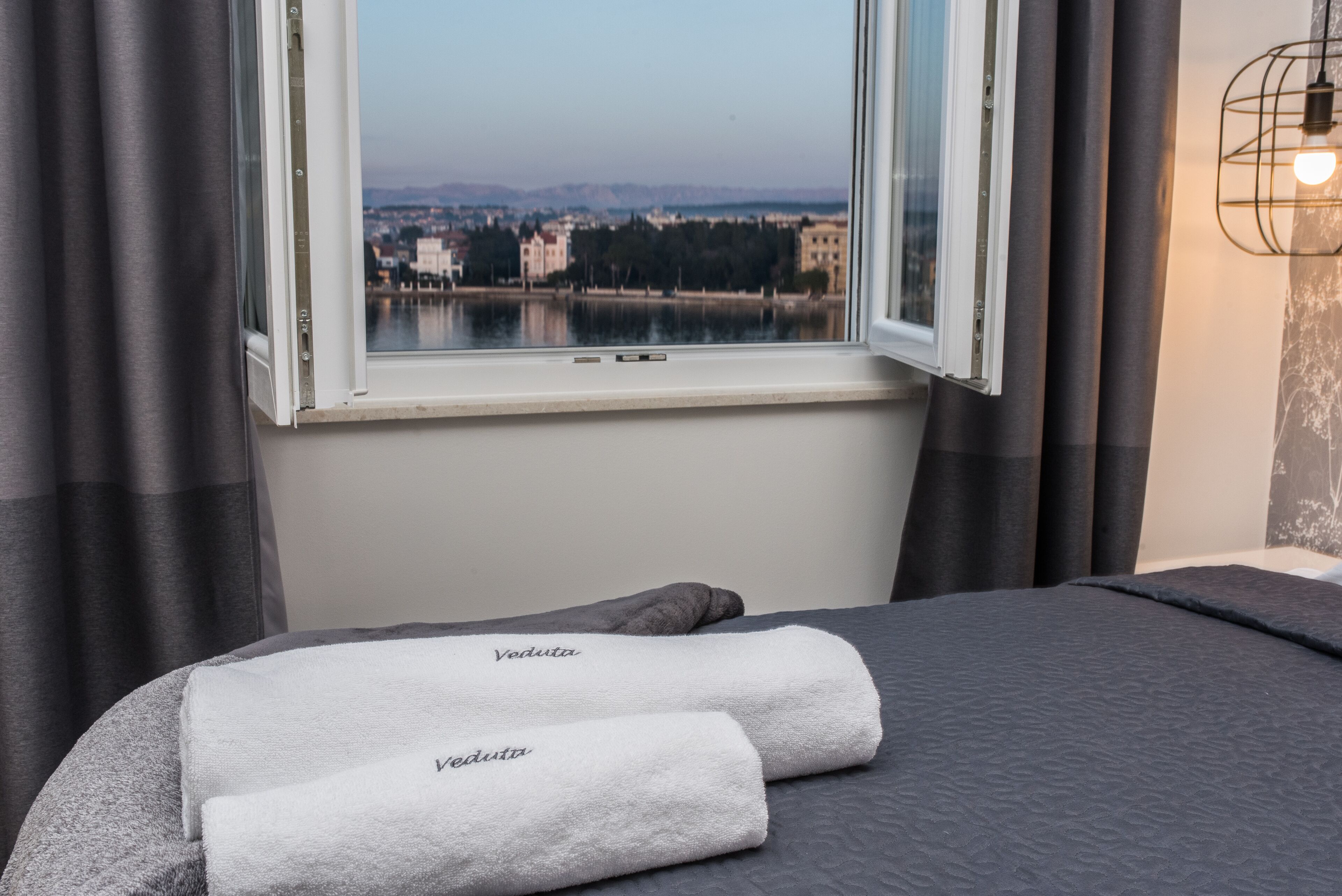 Un lit avec deux serviettes étiquetées « Veduta », et une vue sur un lac et des bâtiments, visible par une fenêtre.