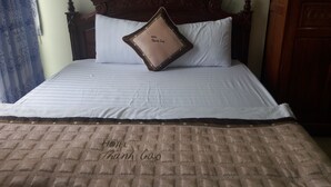 A hotel bed with a quilted bedspread, a pillow with 'HOTEL Thanh Cao' embroidered on it, and a wooden headboard.