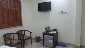 A small room with a television mounted on the wall, a small refrigerator, a wooden desk, two chairs, and a window with wooden shutters.