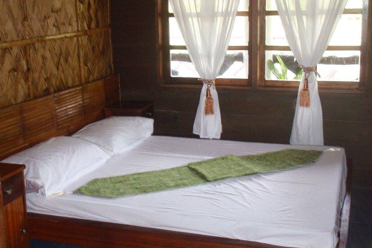 A wooden room with a bed, white curtains, and a window with a view of greenery.