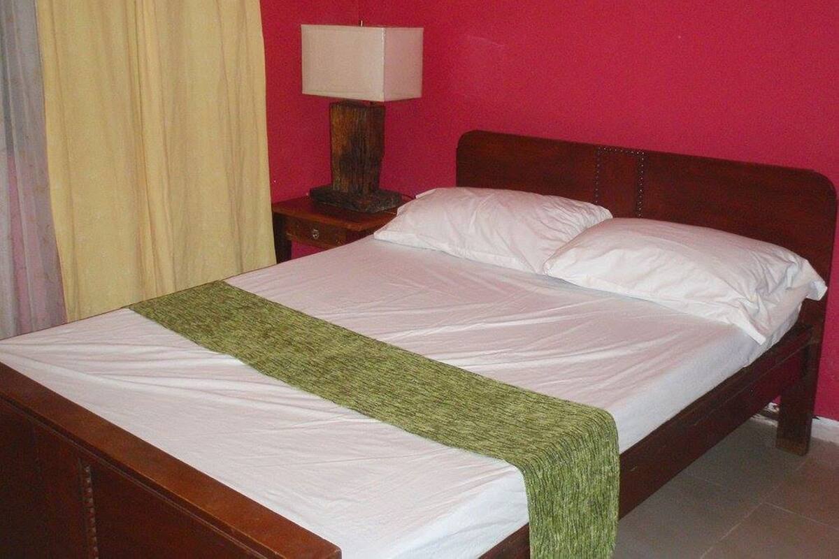 A bed with white linens and a green throw, a wooden nightstand with a lamp, and a pink wall.