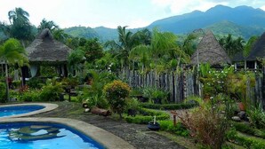 A pool area with thatched-roof structures, lush greenery, and mountains in the background. - Tierra de Lago (Los Banos)