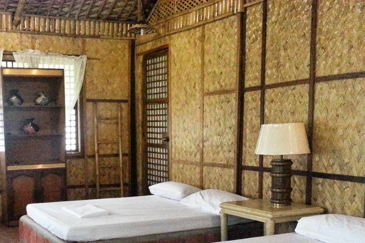 A room with two beds, a wooden table with a lamp, and a window with a decorative screen.