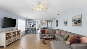 A modern living room with a grey sofa, a television, and a kitchen in the background. - Affordable Renovated 3 Bd/2 Ba home steps from the beach with pool/tennis access (Gulf Shores)