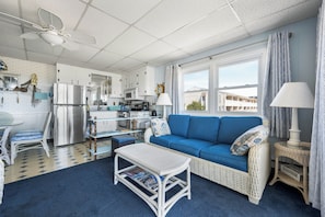 A compact living space with a kitchenette, a blue sofa, a dining area with a table and chairs, and a window with a view of buildings. - Cozy unit located oceanfront on 128th Street! (Ocean City)