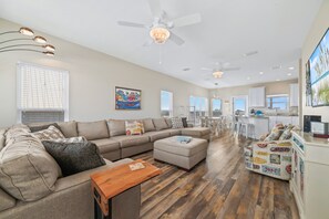 A spacious living room with a large sectional sofa, a wooden coffee table, and a dining area with chairs. There are ceiling fans, recessed lighting, and a kitchen area visible.