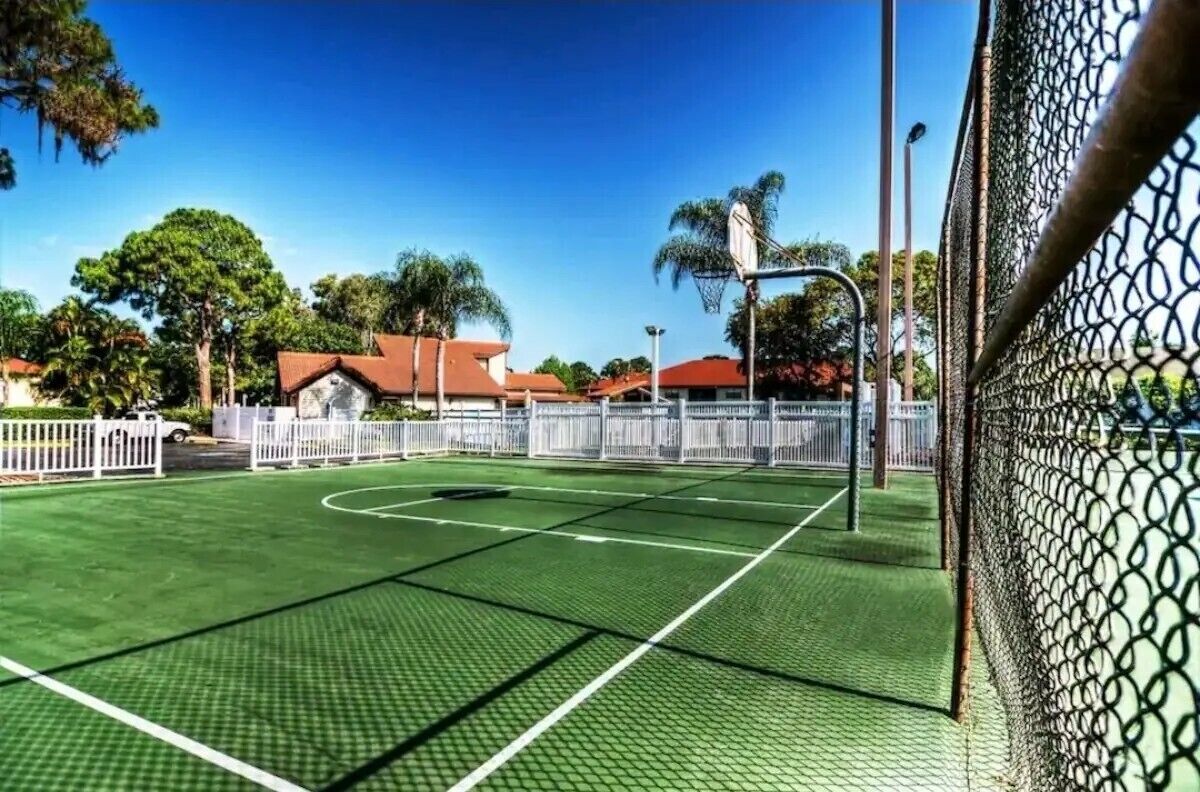 Una cancha de baloncesto con aro, rodeada por una valla blanca y árboles al fondo.