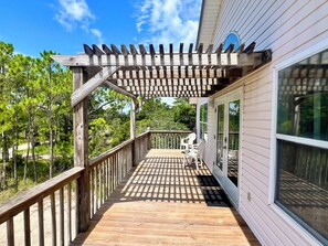 A wooden deck with a pergola, white chairs, and a view of trees.