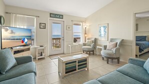 A living room with a television displaying a beach scene, a sign reading "BEACH", and a view of the ocean through a door.