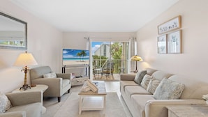A spacious living room with two sofas, a coffee table, and a TV. There is a balcony with a view of palm trees and a house. - Seaside Escape – Beach Castle #24 by RVA (Longboat Key)