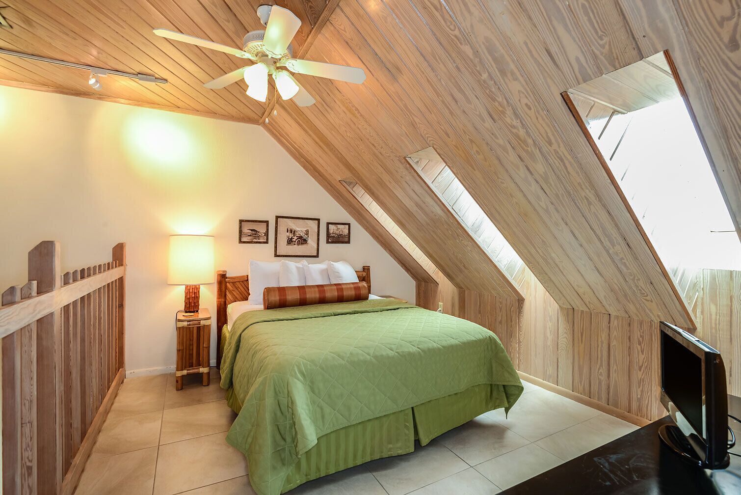 A bedroom with a sloped ceiling, a bed with green bedding, a nightstand with a lamp, and a TV on a stand.