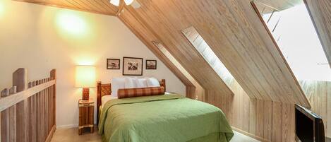 A bedroom with a sloped ceiling, a bed with green bedding, a nightstand with a lamp, and a TV on a stand.