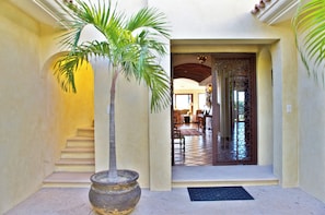 A hallway with a palm tree, a staircase, and a view into a room with a tiled floor and furniture.